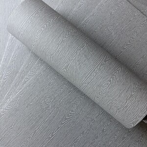 May include: A roll of gray wallpaper with a wood grain pattern. The wallpaper has a textured surface with visible wood knots and grain lines. The roll is partially unrolled, revealing the pattern on the flat surface.