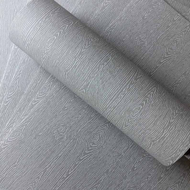 May include: A roll of gray wallpaper with a wood grain pattern. The wallpaper has a textured surface with visible wood knots and grain lines. The roll is partially unrolled, revealing the pattern on the flat surface.