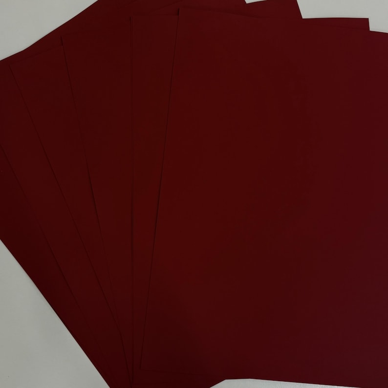 May include: A stack of burgundy-colored cardstock paper fanned out against a white background. The paper has a smooth, matte finish and is evenly cut. The color is a deep, rich shade of red.