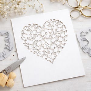 Op de afbeelding: Een witte rechthoekige kaart met een grote, ingewikkelde hartvormige uitsparing met een gedetailleerd bloemen- en bladpatroon. De kaart ligt naast een metalen schaar, kleine stukjes kurk en gedroogde witte bloemen op een lichte ondergrond.