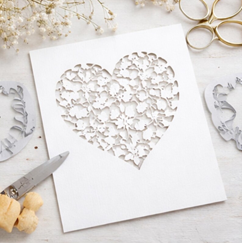 Op de afbeelding: Een witte rechthoekige kaart met een grote, ingewikkelde hartvormige uitsparing met een gedetailleerd bloemen- en bladpatroon. De kaart ligt naast een metalen schaar, kleine stukjes kurk en gedroogde witte bloemen op een lichte ondergrond.