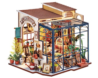 Pode incluir: Modelo em miniatura de uma floricultura com estufa. O modelo apresenta um telhado castanho, paredes bege e uma estufa de vidro com moldura azul. A placa diz "FLOWERSHOP". O interior é preenchido com plantas em miniatura, móveis e objetos decorativos.