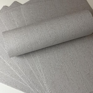 May include: Gray wood grain wallpaper, with a roll of wallpaper in the foreground. The wallpaper has a detailed wood grain pattern with knots and vertical lines, creating a realistic wood effect.