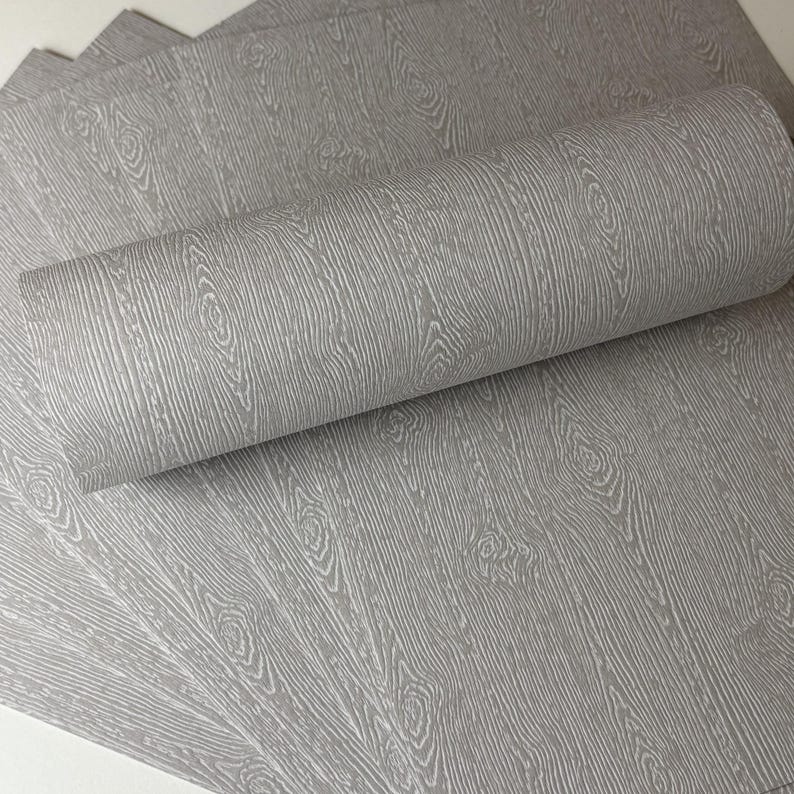 May include: Gray wood grain wallpaper, with a roll of wallpaper in the foreground. The wallpaper has a detailed wood grain pattern with knots and vertical lines, creating a realistic wood effect.