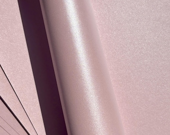 May include: Close-up of a roll of light pink, shimmering paper. The paper has a subtle glitter effect and a smooth, reflective surface. Several sheets of the same paper are visible in the lower left corner, with a shadow cast across the surface.