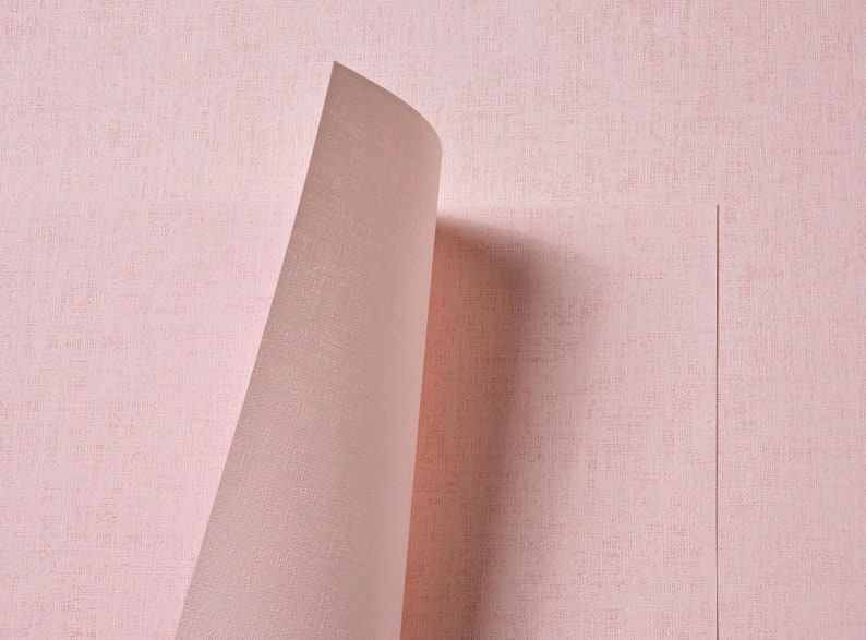 May include: A roll of light pink wallpaper with a subtle, woven fabric-like texture. The paper is partially unrolled, revealing a curved edge that casts a soft shadow onto the flat, vertical section of the same pink, textured material.