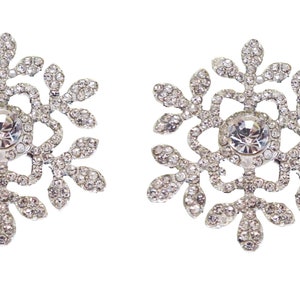 May include: A pair of silver-coloured snowflake brooches. Each brooch has a central, large, clear crystal surrounded by smaller crystals. The snowflake design has six points with leaf-like details, all encrusted with sparkling crystals.