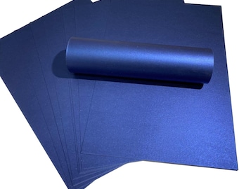 May include: A stack of navy blue paper sheets with a metallic sheen, alongside a rolled-up tube of matching paper. The paper has a smooth, reflective surface. The items are arranged on a white background, suggesting a stationery or craft supply.