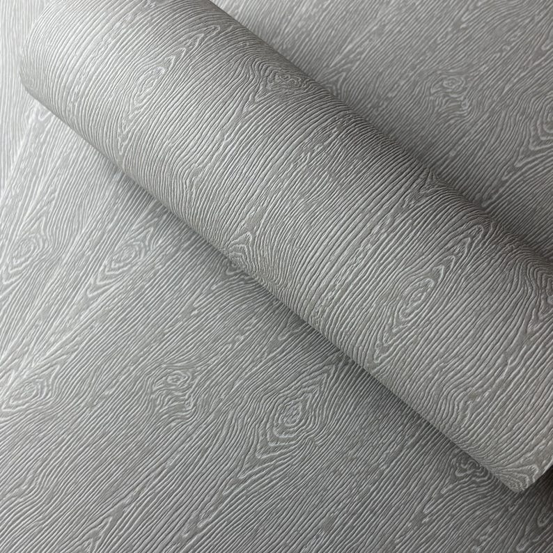 May include: A roll of gray wallpaper with a wood grain pattern. The wallpaper has a textured surface with visible wood grain lines and knots. The color is a neutral gray, suitable for various interior design styles.