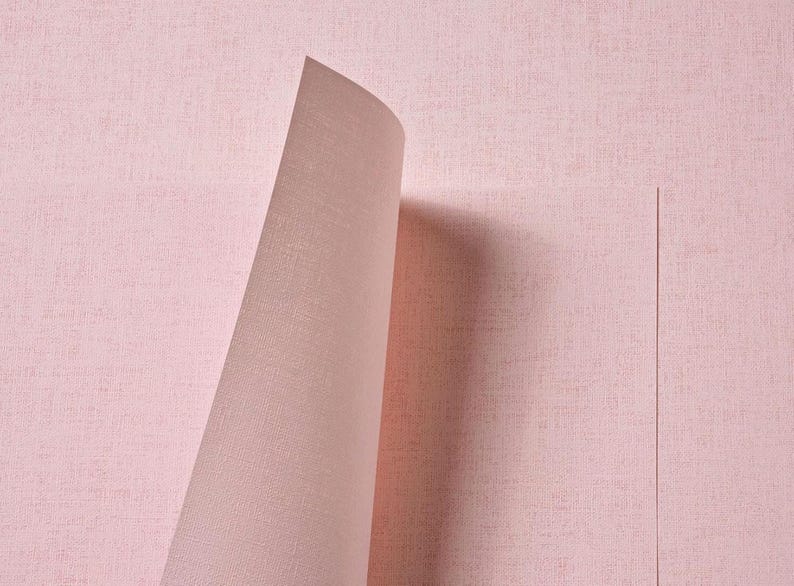 May include: A roll of light pink wallpaper featuring a subtle, woven fabric-like texture. The material is shown partially unrolled, revealing a smooth, consistent surface with a matte finish and a fine, cross-hatched pattern across the paper.