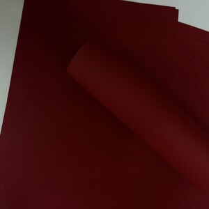 May include: A stack of deep red sheets of paper, with one sheet rolled up. The paper has a smooth, matte finish. The color is a rich, dark shade of red, similar to burgundy or maroon. The paper is likely for crafts or stationery.