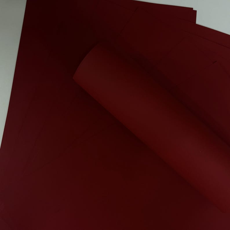 May include: A stack of deep red sheets of paper, with one sheet rolled up. The paper has a smooth, matte finish. The color is a rich, dark shade of red, similar to burgundy or maroon. The paper is likely for crafts or stationery.