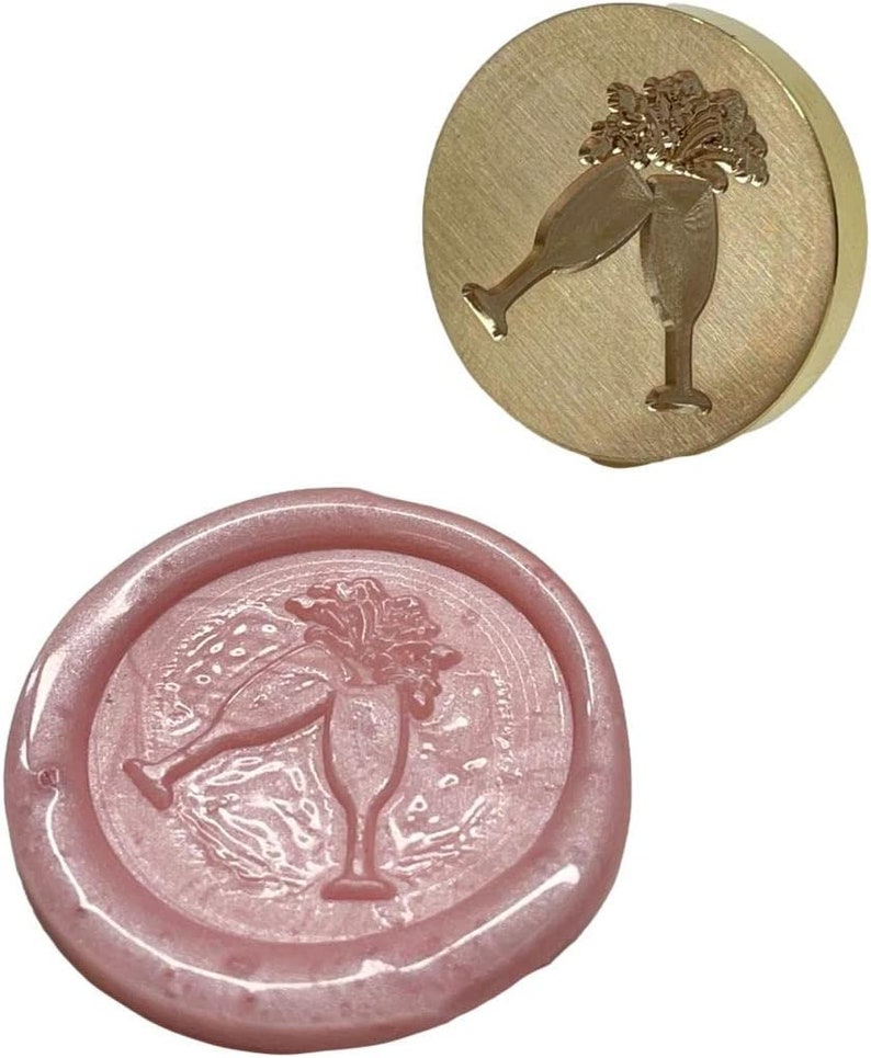 May include: A gold-toned metal wax seal stamp with a design of two champagne glasses clinking together. The stamp is used to create a wax seal with the same design.