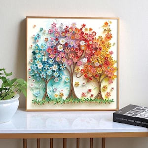 May include: A framed artwork featuring three stylized trees with colorful, paper-like flowers. The trees transition from blue to red to yellow, with brown trunks and green grass at the base. The artwork is displayed on a white surface.