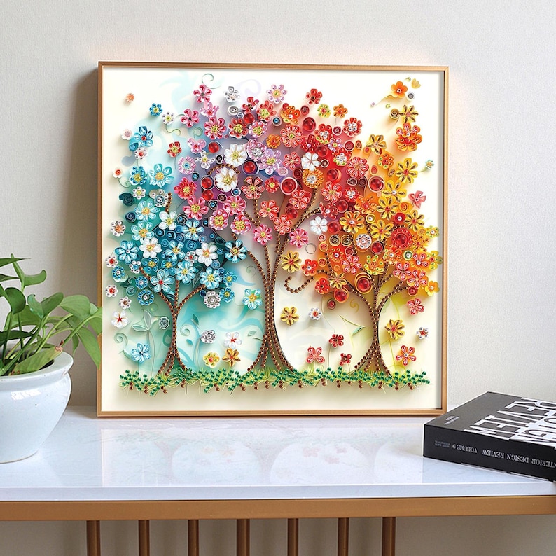 May include: A framed artwork featuring three stylized trees with colorful, paper-like flowers. The trees transition from blue to red to yellow, with brown trunks and green grass at the base. The artwork is displayed on a white surface.