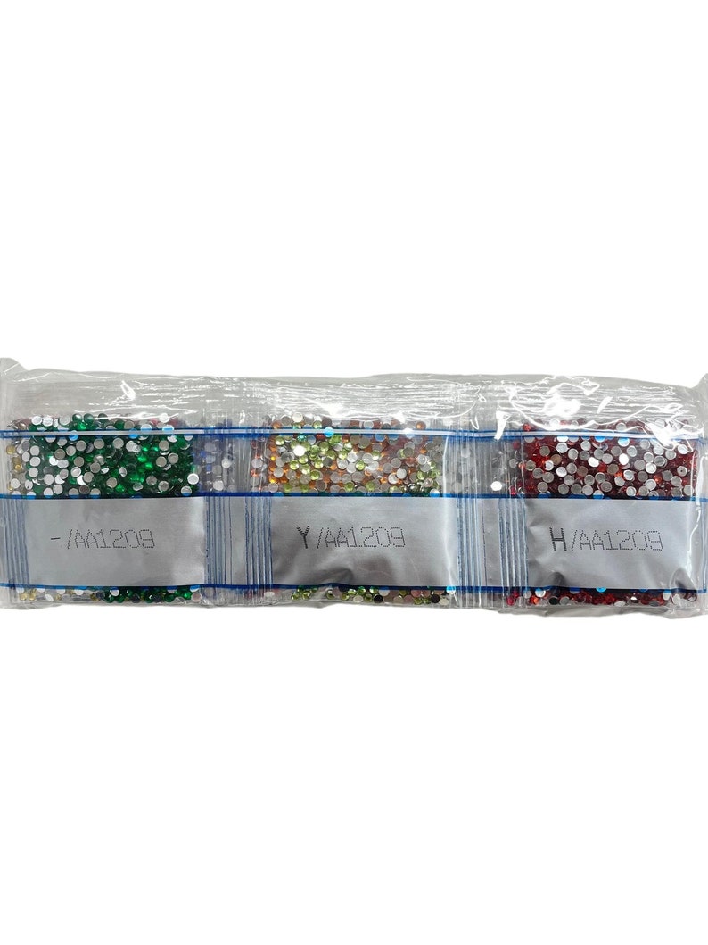 May include: Three small, clear plastic bags filled with colorful, sparkling rhinestones. The bags are labeled with letters and numbers. The rhinestones are in shades of green, gold, and red. The bags are sealed together.