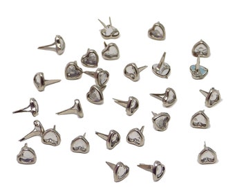 May include: A collection of silver-colored metal heart-shaped studs with clear, faceted centers. The studs have a pointed post on the back for attachment. The studs are scattered on a white background.