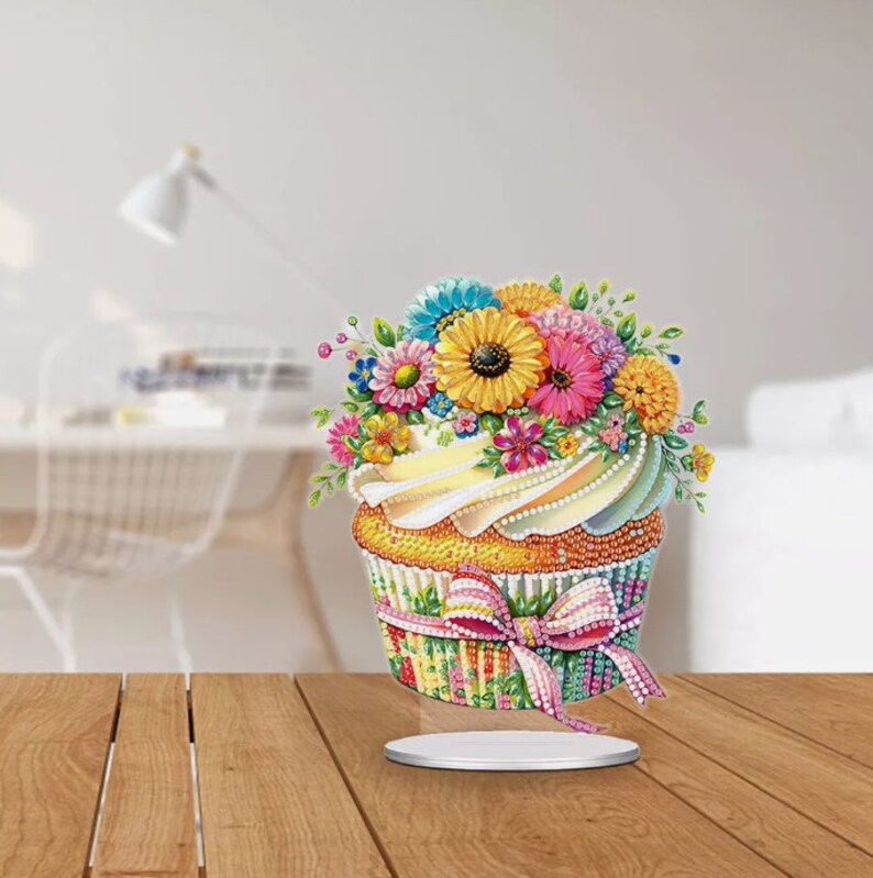 May include: A colorful cupcake with a white frosting and a bouquet of flowers on top. The cupcake is decorated with a pink ribbon and a striped pattern.
