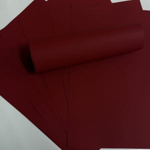 May include: A stack of burgundy-colored paper sheets with a rolled-up sheet on top. The paper has a smooth texture and a deep, rich color. The sheets are fanned out, showing the edges of the paper.