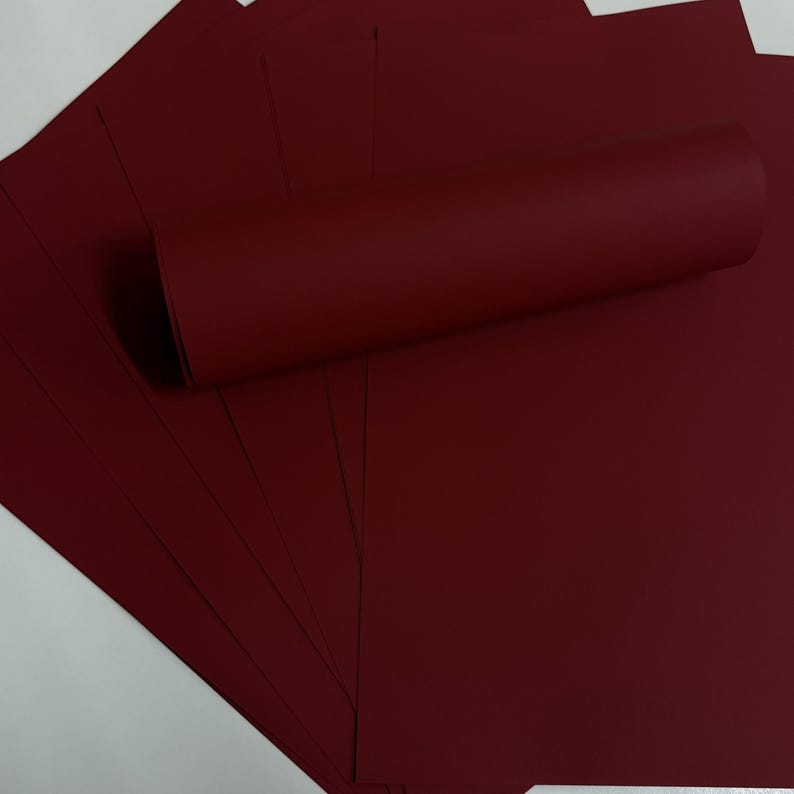 May include: A stack of burgundy-colored paper sheets with a rolled-up sheet on top. The paper has a smooth texture and a deep, rich color. The sheets are fanned out, showing the edges of the paper.