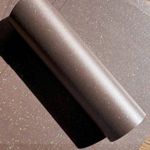 May include: A roll of brown glitter vinyl with a shimmering, iridescent effect. The vinyl is rolled up, with a portion unrolled, revealing the glittery surface. The background is a matching sheet of glitter vinyl.