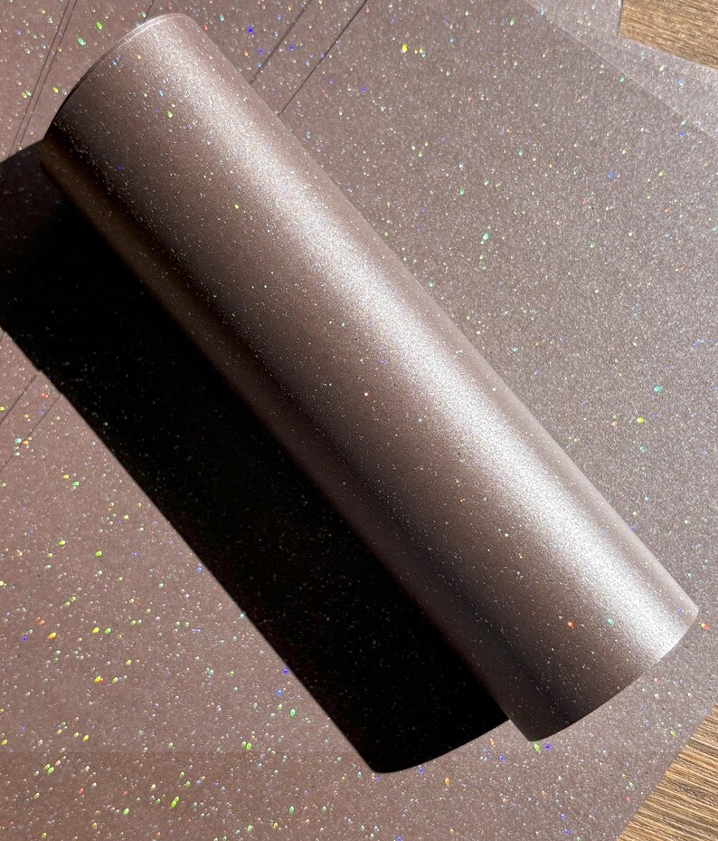 May include: A roll of brown glitter vinyl with a shimmering, iridescent effect. The vinyl is rolled up, with a portion unrolled, revealing the glittery surface. The background is a matching sheet of glitter vinyl.