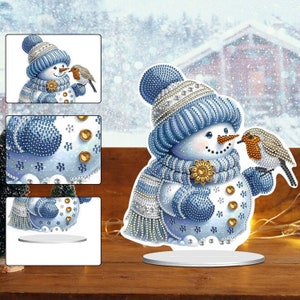 May include: A blue and white snowman with a red nose and a yellow flower on its chest. A small brown and white bird is perched on the snowman's arm. The snowman is standing on a white base.