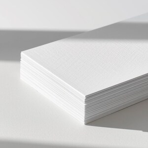 Op de afbeelding: Een stapel rechthoekige witte kaarten met een zichtbare textuurafwerking. De kaarten zijn netjes opgestapeld, waarbij de dikte van het papier en het fijne korrelpatroon op het oppervlak van elk vel wit karton duidelijk zichtbaar zijn.