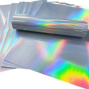 May include: A stack of iridescent holographic vinyl sheets in a silver color with a rainbow sheen. The sheets are rolled up and stacked on top of each other.