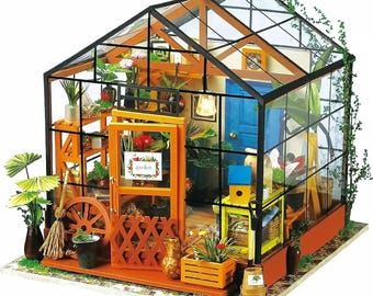May include: A miniature greenhouse model with a glass roof and walls, featuring a wooden frame and interior details. The model includes miniature plants, a blue door, and a sign that reads "garden".