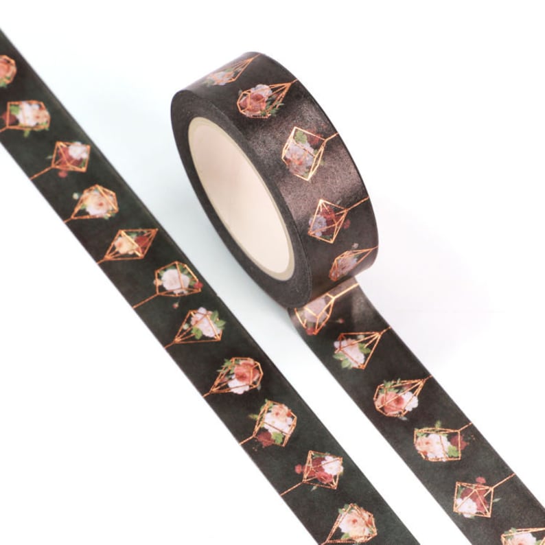 May include: A roll of black and gold washi tape with a floral pattern. The tape features a repeating design of gold geometric shapes with pink and white flowers inside.