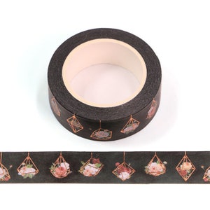 May include: A roll of black washi tape with a gold geometric design and floral print. The tape features a repeating pattern of hanging geometric shapes with floral arrangements inside.