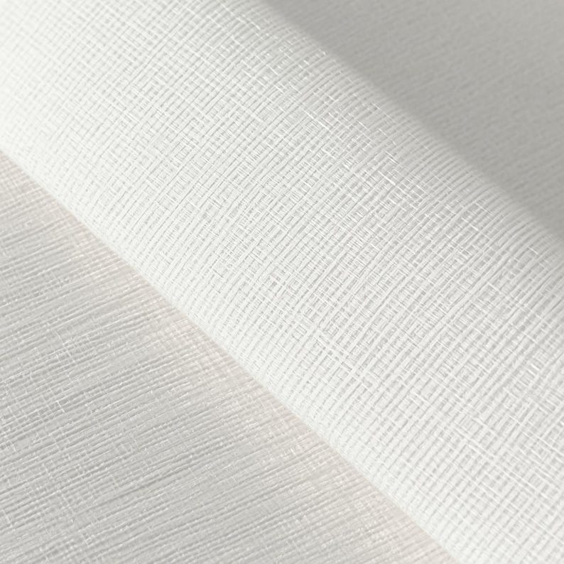 Op de afbeelding: Een rol witte stof met een duidelijke kruislings gearceerde textuur. Het materiaal vertoont een rasterachtig patroon van fijne, elkaar kruisende lijnen die een subtiel, verhoogd oppervlakte-effect cre&euml;ren over het gehele witte oppervlak.
