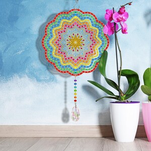 May include: A colorful mandala-shaped wall hanging with a rainbow crystal pendant. The mandala is made of colorful beads and has a starburst design in the center. The hanging is displayed against a blue wall with a potted orchid plant.
