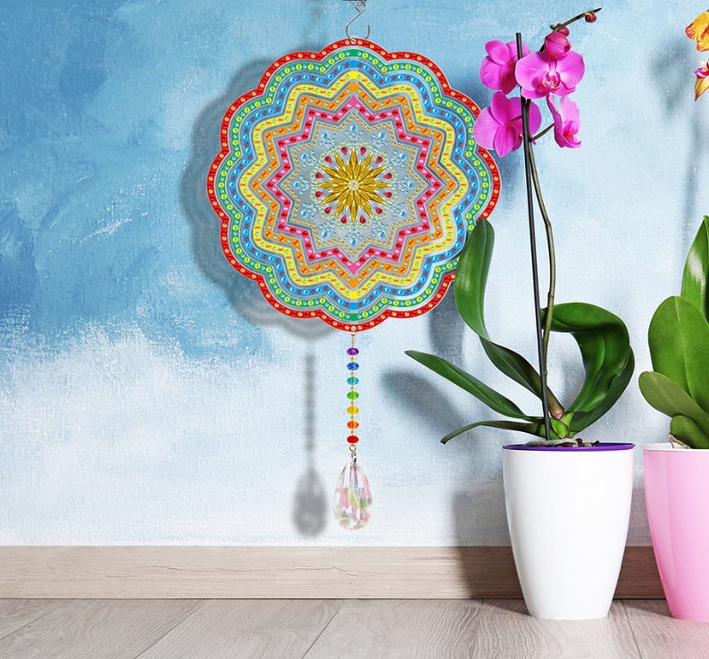 May include: A colorful mandala-shaped wall hanging with a rainbow crystal pendant. The mandala is made of colorful beads and has a starburst design in the center. The hanging is displayed against a blue wall with a potted orchid plant.