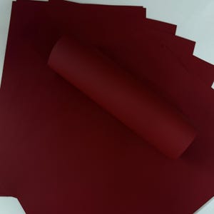 May include: A stack of burgundy-colored paper sheets with a rolled-up sheet on top. The paper has a smooth texture and a deep, rich color. The background is white.