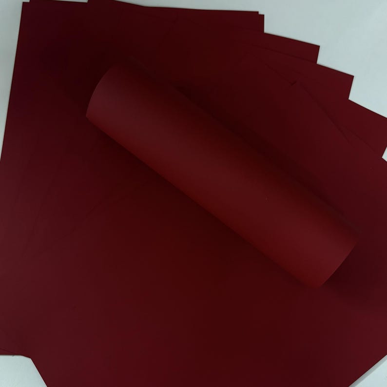 May include: A stack of burgundy-colored paper sheets with a rolled-up sheet on top. The paper has a smooth texture and a deep, rich color. The background is white.