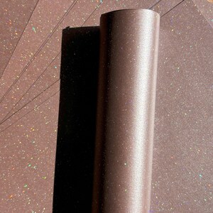 May include: A brown, cylindrical object stands upright, casting a dark shadow on a fan of brown, glitter-speckled paper. The paper and cylinder have a subtle sheen, with iridescent glitter adding a touch of sparkle. The image is well-lit, highlighting the textures.
