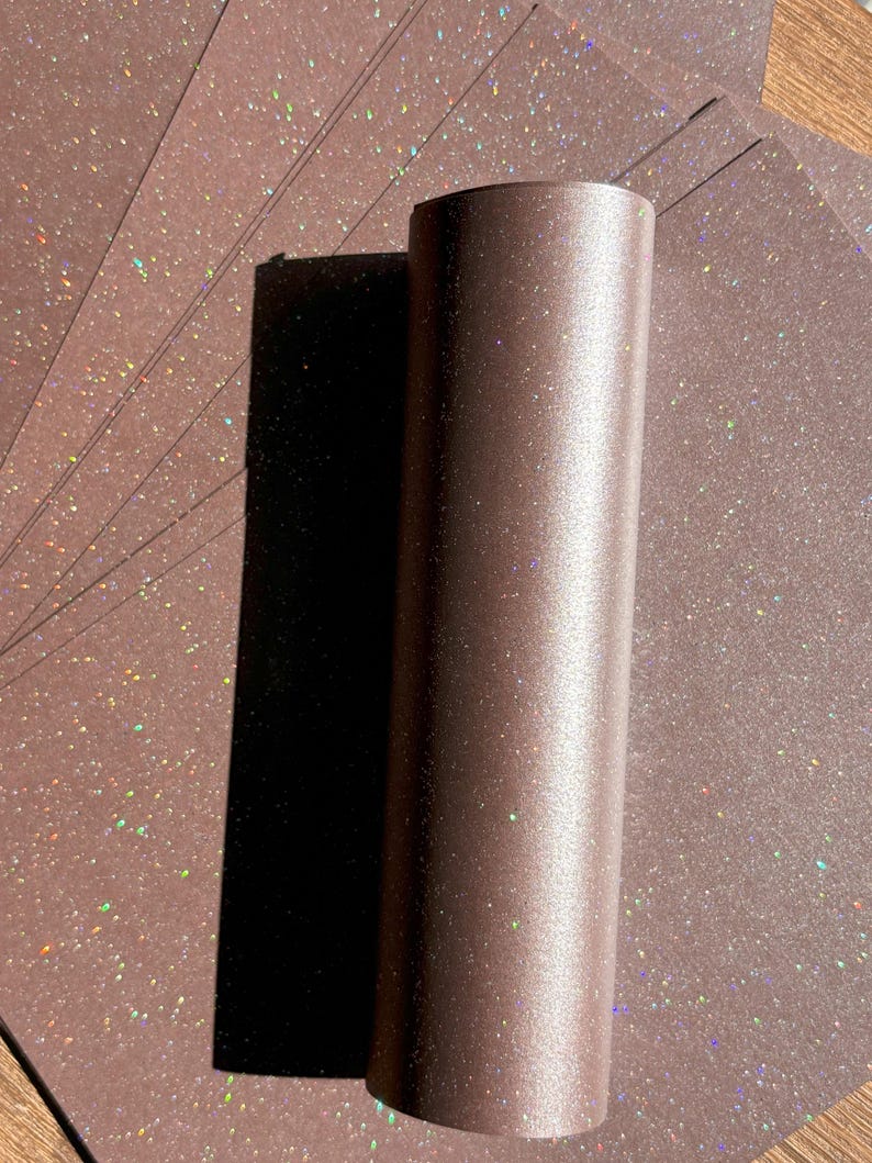 May include: A brown, cylindrical object stands upright, casting a dark shadow on a fan of brown, glitter-speckled paper. The paper and cylinder have a subtle sheen, with iridescent glitter adding a touch of sparkle. The image is well-lit, highlighting the textures.