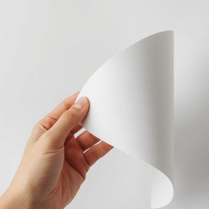 Op de afbeelding: Een flexibel, wit, getextureerd vel papier of canvasmateriaal dat in een gebogen positie wordt gehouden. Het oppervlak vertoont een fijn, uniform korrelpatroon over het hele gebied, met een heldere witte kleur en een gladde, matte afwerking.