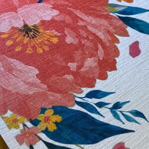 Op de afbeelding: Een gestructureerd wit oppervlak met een grote, levendige koraalroze pioenroosillustratie met een geel hart, geaccentueerd door kleinere gele bloemen en donkergroene bladeren in petrol. Het bloemmotief heeft een zichtbaar textielachtig patroon.