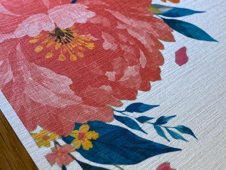 Op de afbeelding: Een gestructureerd wit oppervlak met een grote, levendige koraalroze pioenroosillustratie met een geel hart, geaccentueerd door kleinere gele bloemen en donkergroene bladeren in petrol. Het bloemmotief heeft een zichtbaar textielachtig patroon.