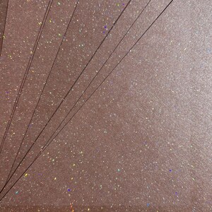 May include: A close-up of multiple sheets of rose-gold glitter paper. The paper has a textured surface with iridescent specks. Thin, dark lines are visible. The glitter catches the light, creating a shimmering effect. The overall aesthetic is elegant and festive.