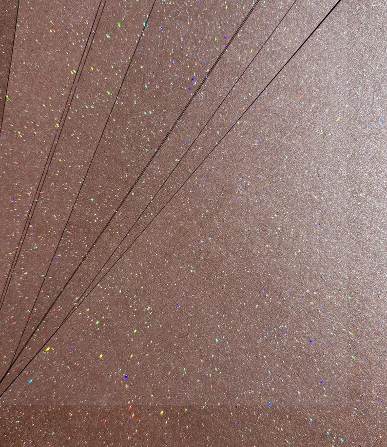 May include: A close-up of multiple sheets of rose-gold glitter paper. The paper has a textured surface with iridescent specks. Thin, dark lines are visible. The glitter catches the light, creating a shimmering effect. The overall aesthetic is elegant and festive.