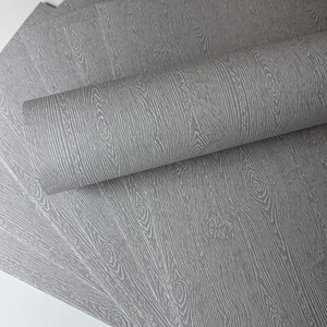 May include: Gray wood grain wallpaper, partially rolled, with a detailed pattern of vertical lines and circular knots. The wallpaper has a textured appearance, creating a realistic wood effect. Several sheets are visible.
