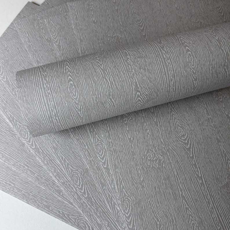 May include: Gray wood grain wallpaper, partially rolled, with a detailed pattern of vertical lines and circular knots. The wallpaper has a textured appearance, creating a realistic wood effect. Several sheets are visible.
