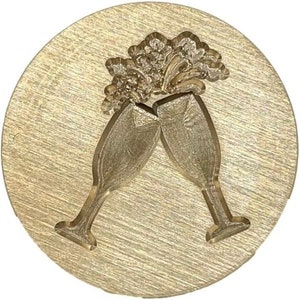 May include: A gold-toned metal stamp with a design of two champagne flutes clinking together. The flutes are raised in a toast, with a stylized design of bubbles above them.