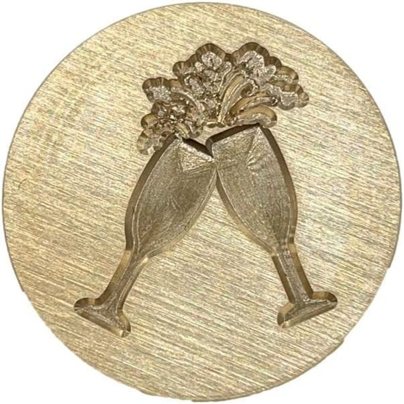May include: A gold-toned metal stamp with a design of two champagne flutes clinking together. The flutes are raised in a toast, with a stylized design of bubbles above them.