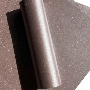 May include: A roll of brown glitter paper with a metallic sheen, partially unrolled to reveal individual sheets. The paper has a sparkling, iridescent effect, catching the light with flecks of colour. Ideal for crafts and scrapbooking.