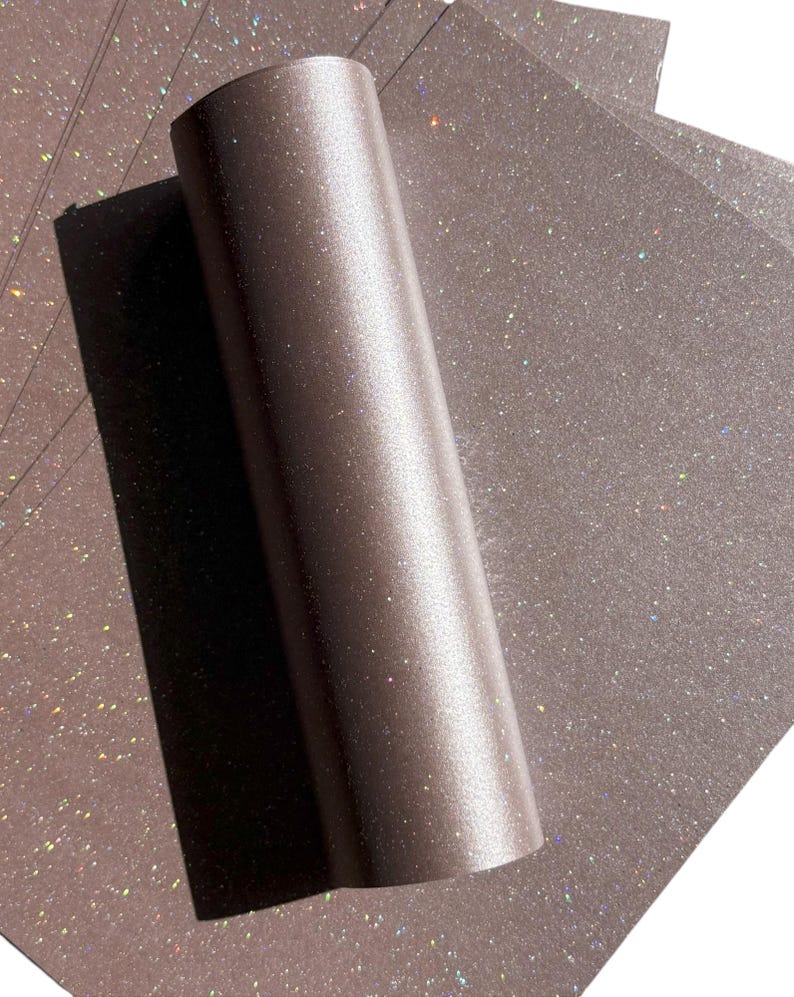 May include: A roll of brown glitter paper with a metallic sheen, partially unrolled to reveal individual sheets. The paper has a sparkling, iridescent effect, catching the light with flecks of colour. Ideal for crafts and scrapbooking.